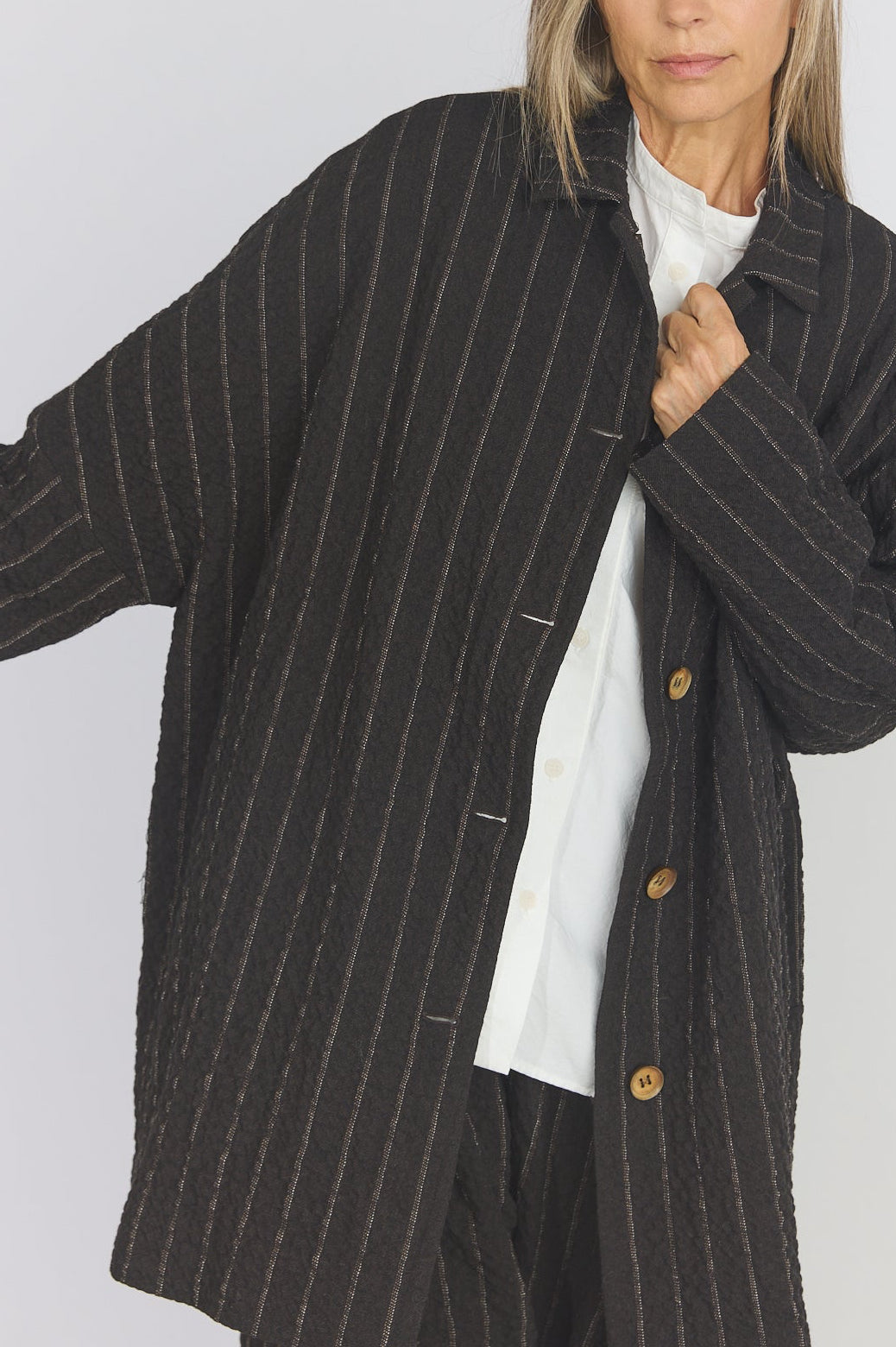 A woman modeling a dark charcoal pinstriped button-up jacket over a white t-shirt, photographed against a plain light gray studio background.