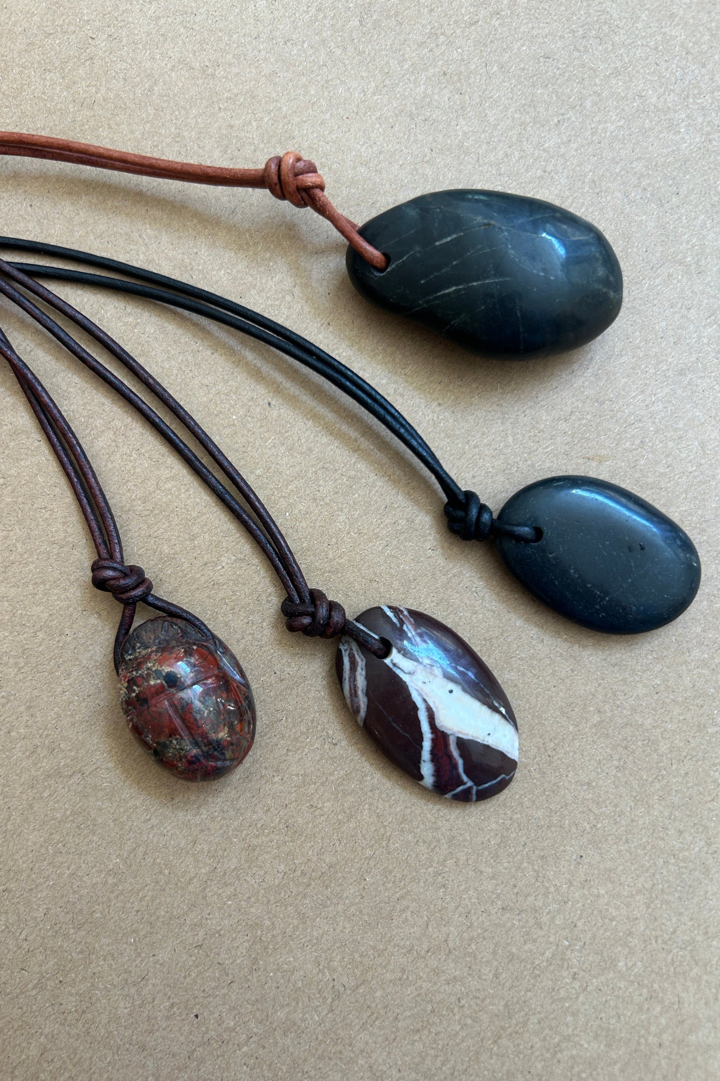 Four polished stone pendants on cords, arranged on a beige textured background. Two pendants are dark charcoal/black, one is a brown-and-white marbled stone, and one is a reddish-brown patterned stone.