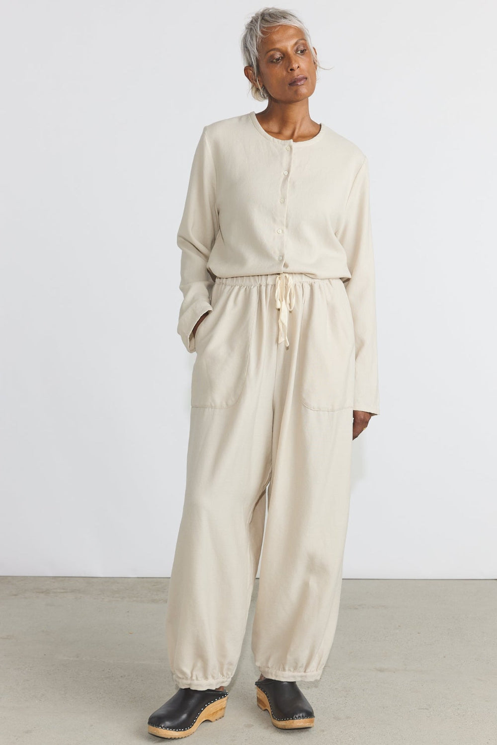 A model wearing a light beige linen jumpsuit with a drawstring waist, long sleeves, and wide-leg pants, paired with black clogs, in a minimalist studio setting.