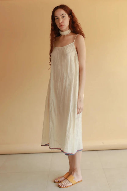 A woman with long wavy red hair stands indoors wearing a loose, sleeveless cream-colored midi dress with thin straps and a small multicolor striped border at the hem, paired with tan slide sandals.