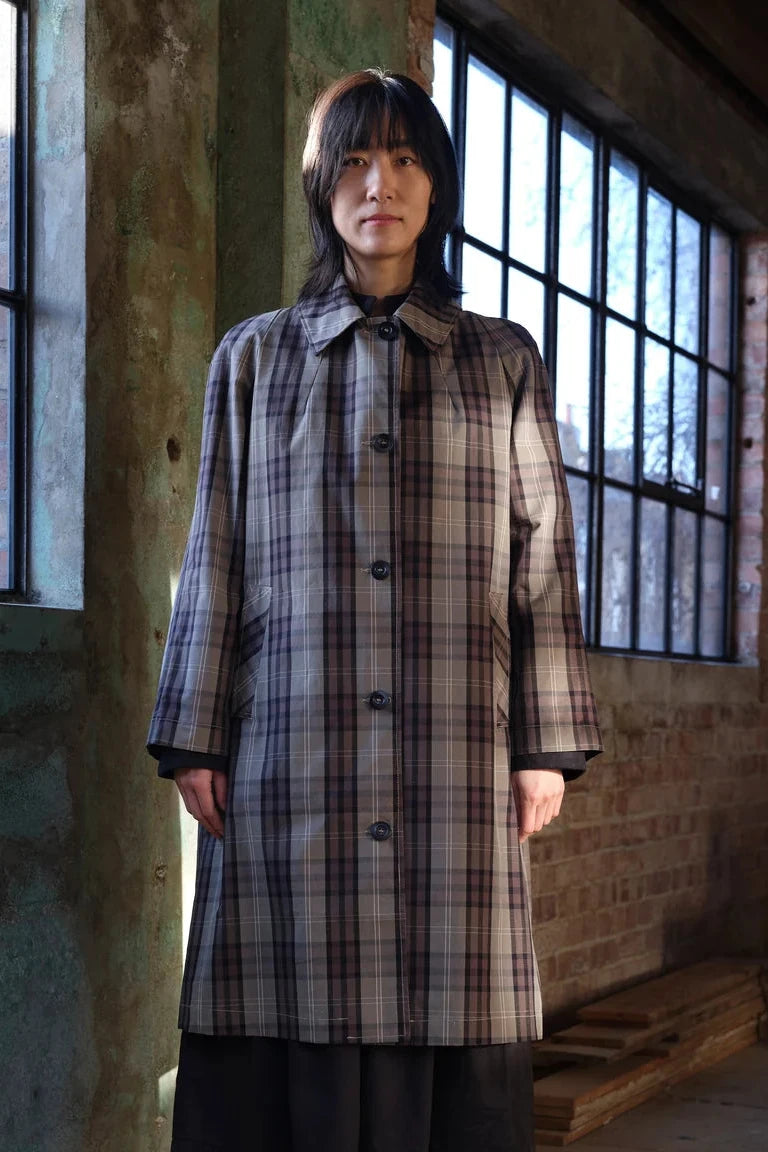A person stands indoors wearing a long plaid coat in muted beige, gray, and brown tones, with dark bottoms. The setting is a rustic room with large black-framed windows, exposed brick walls, and a wooden bench.