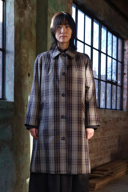 A person stands indoors wearing a long plaid coat in muted beige, gray, and brown tones, with dark bottoms. The setting is a rustic room with large black-framed windows, exposed brick walls, and a wooden bench.