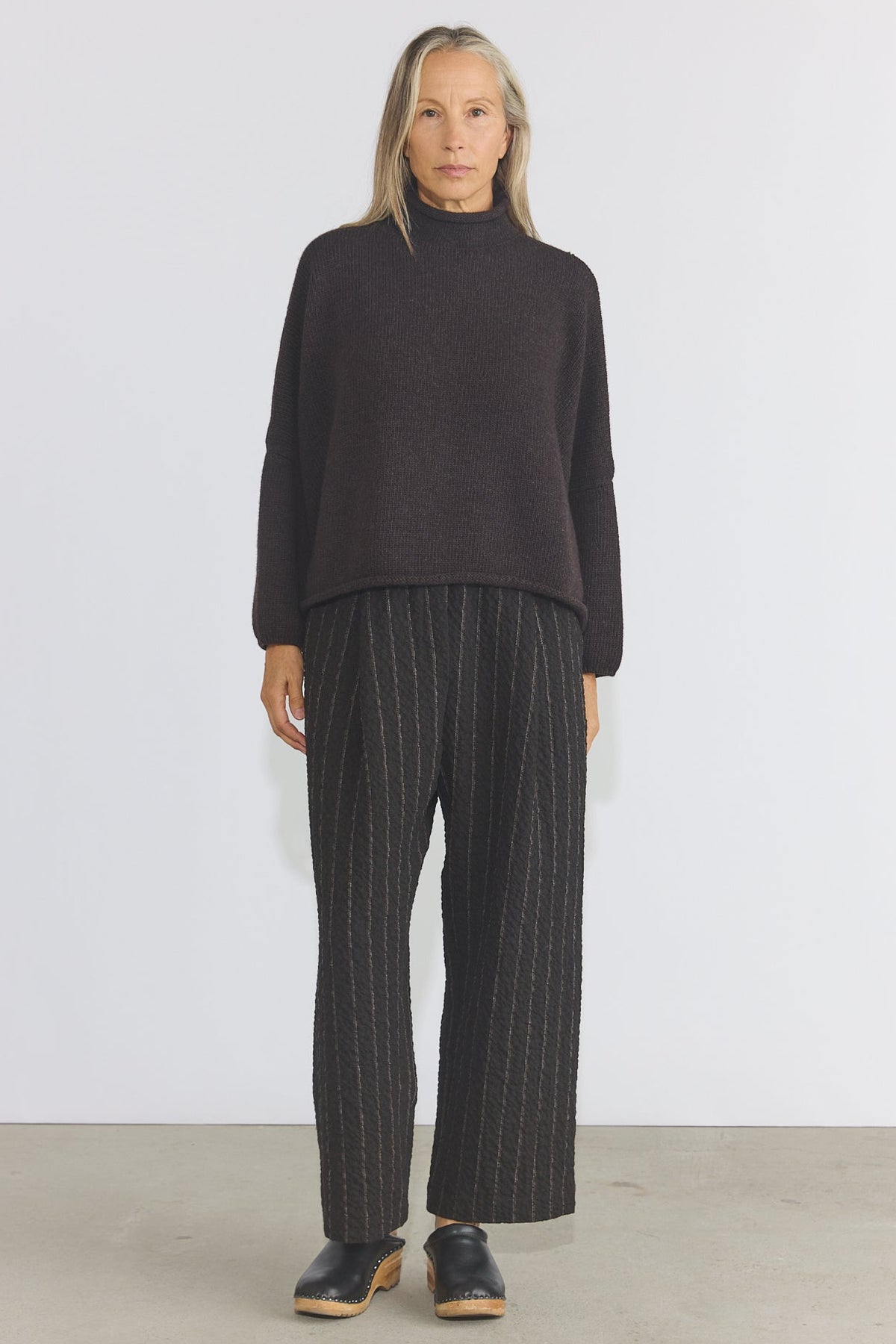 A woman stands facing forward in a studio wearing a dark charcoal turtleneck sweater and dark wide-leg trousers with thin light pinstripes, paired with dark clogs.
