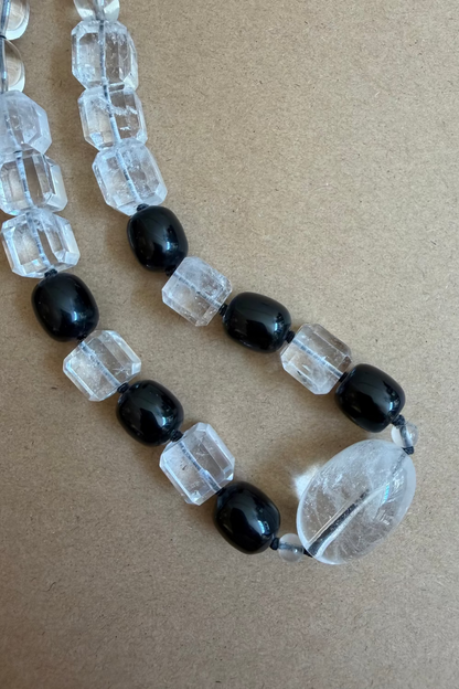 A beaded strand necklace featuring glossy black oval beads and faceted clear crystal beads, with a large clear teardrop-shaped bead at the end, laid on a brown textured background.