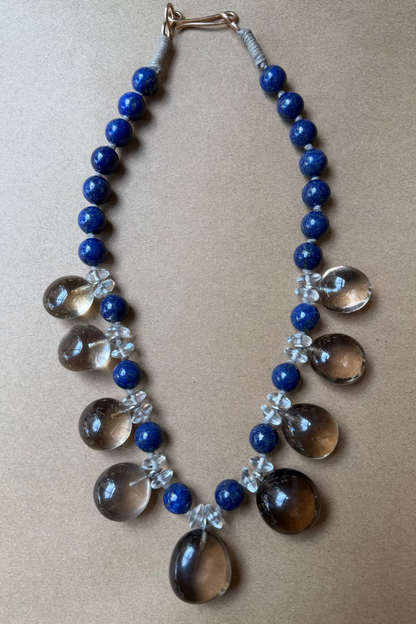 A beaded necklace featuring deep blue round beads, clear teardrop-shaped smoky quartz pendants, and small clear crystal flower motifs, displayed on a neutral tan background.