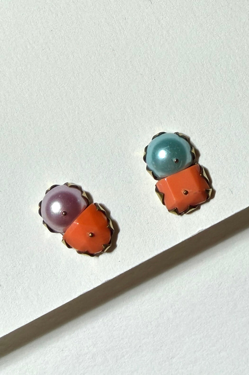 A pair of gold-tone stud earrings with a split two-tone design, featuring a smooth oval purple stone on one side and a textured orange stone on the other, displayed on a white textured surface.