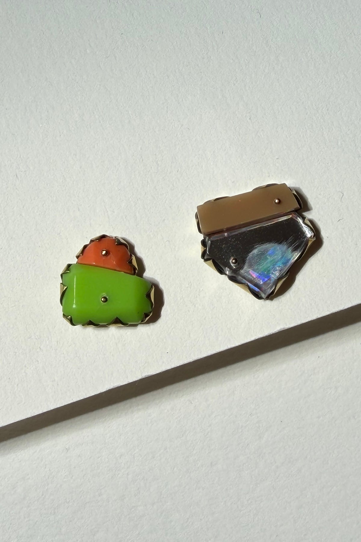 Two small handmade jewelry pieces, likely enamel pins or charms, displayed on a textured off-white background with a wooden stick for scale. One piece features stacked green and orange shapes, and the other has a brown top and a blue-green iridescent center.