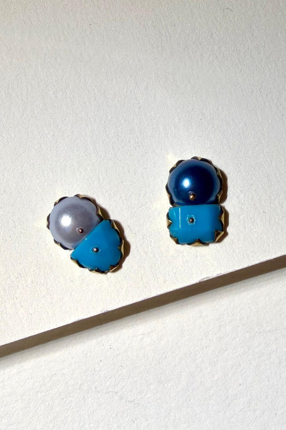 A pair of vintage-style gold-tone stud earrings with blue enamel and pearl accents, displayed on a textured off-white surface with a diagonal shadow.
