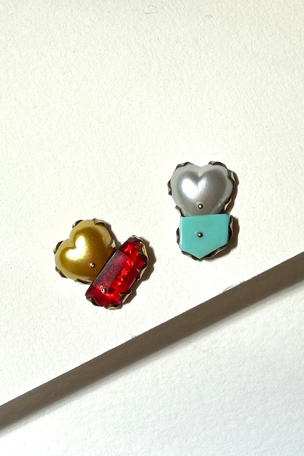 Three small heart-shaped enamel pins with gold-tone borders, displayed on a white surface with a diagonal shadow. The pins feature gold, silver, red, and turquoise colors.