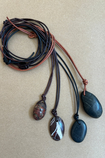 Four polished stone pendants on multi-colored cords, displayed on a textured beige background.