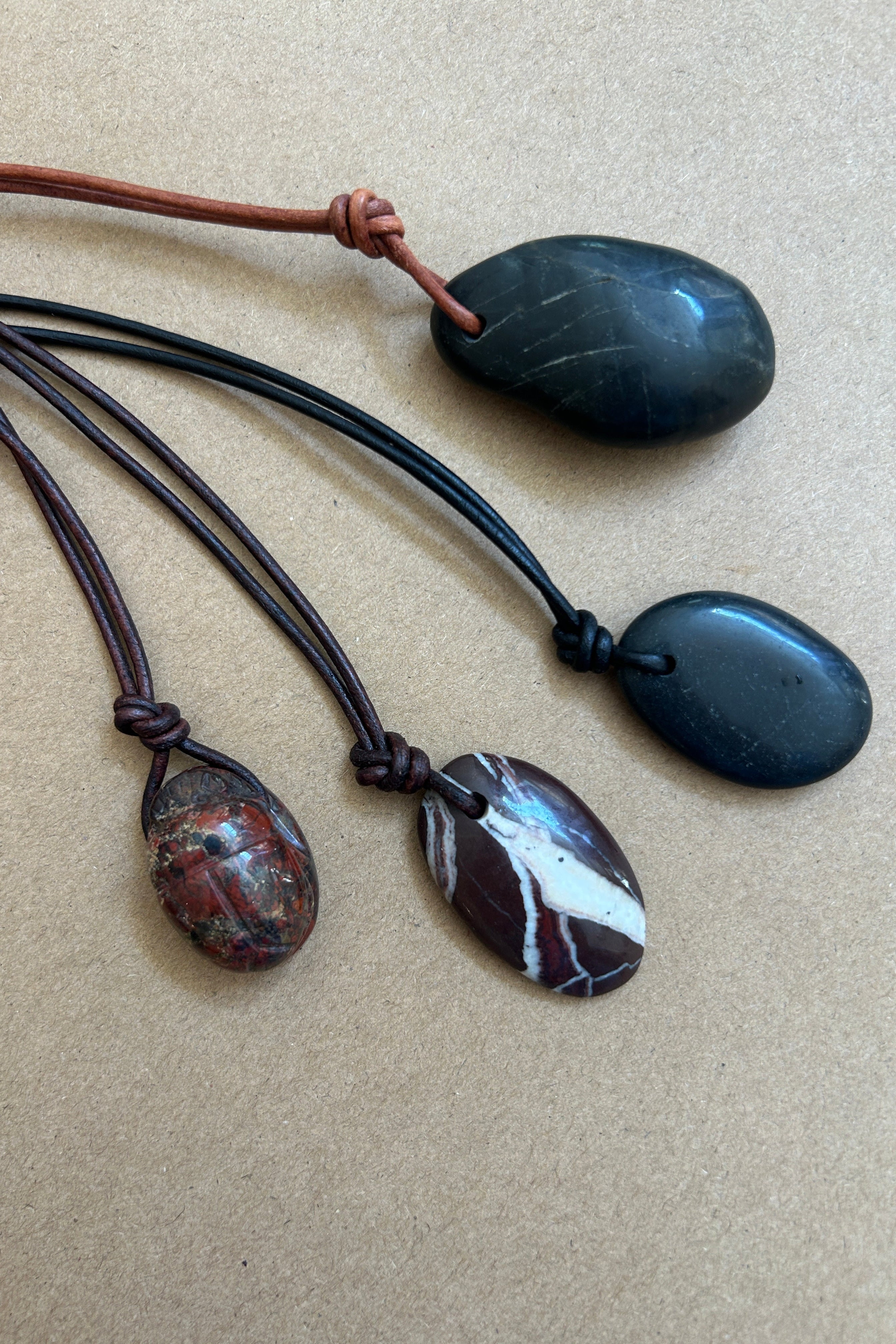 Four polished stone pendants on cords, arranged on a beige textured background. Two pendants are dark charcoal/black, one is a brown-and-white marbled stone, and one is a reddish-brown patterned stone.