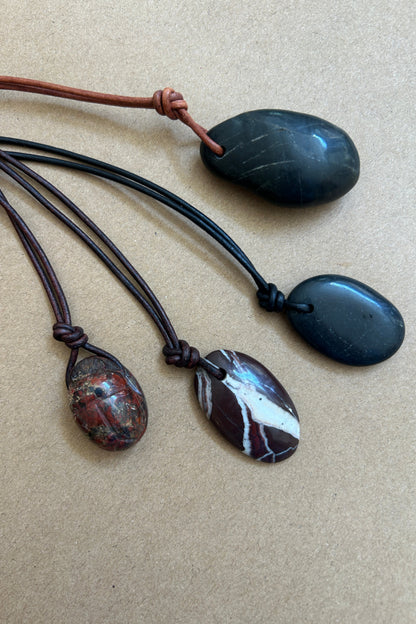 Four polished stone pendants on cords, arranged on a beige textured background. Two pendants are dark charcoal/black, one is a brown-and-white marbled stone, and one is a reddish-brown patterned stone.