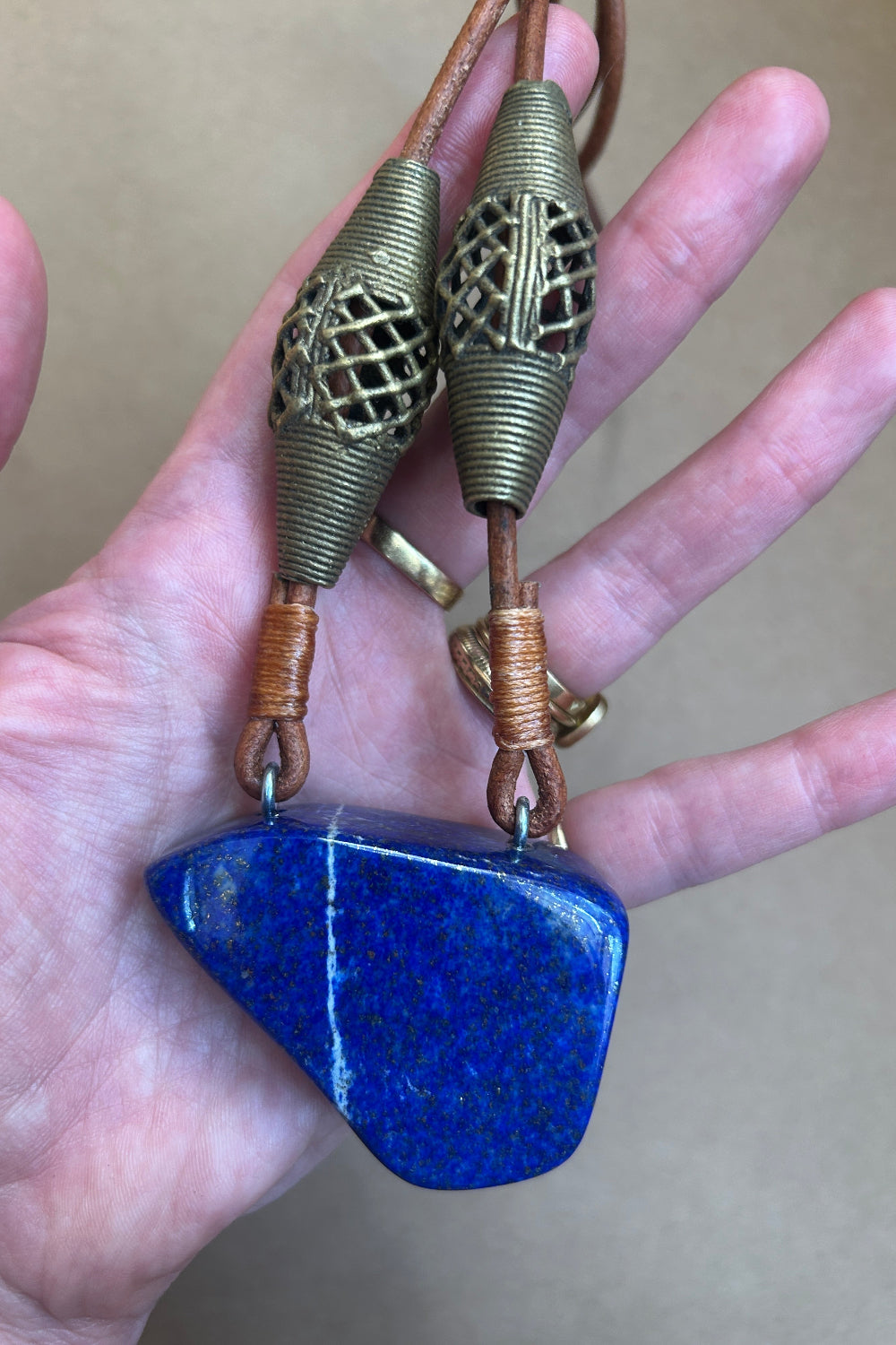A hand holding a pendant necklace featuring a polished blue lapis lazuli stone and two bronze-colored wire-wrapped beads on a brown cord.