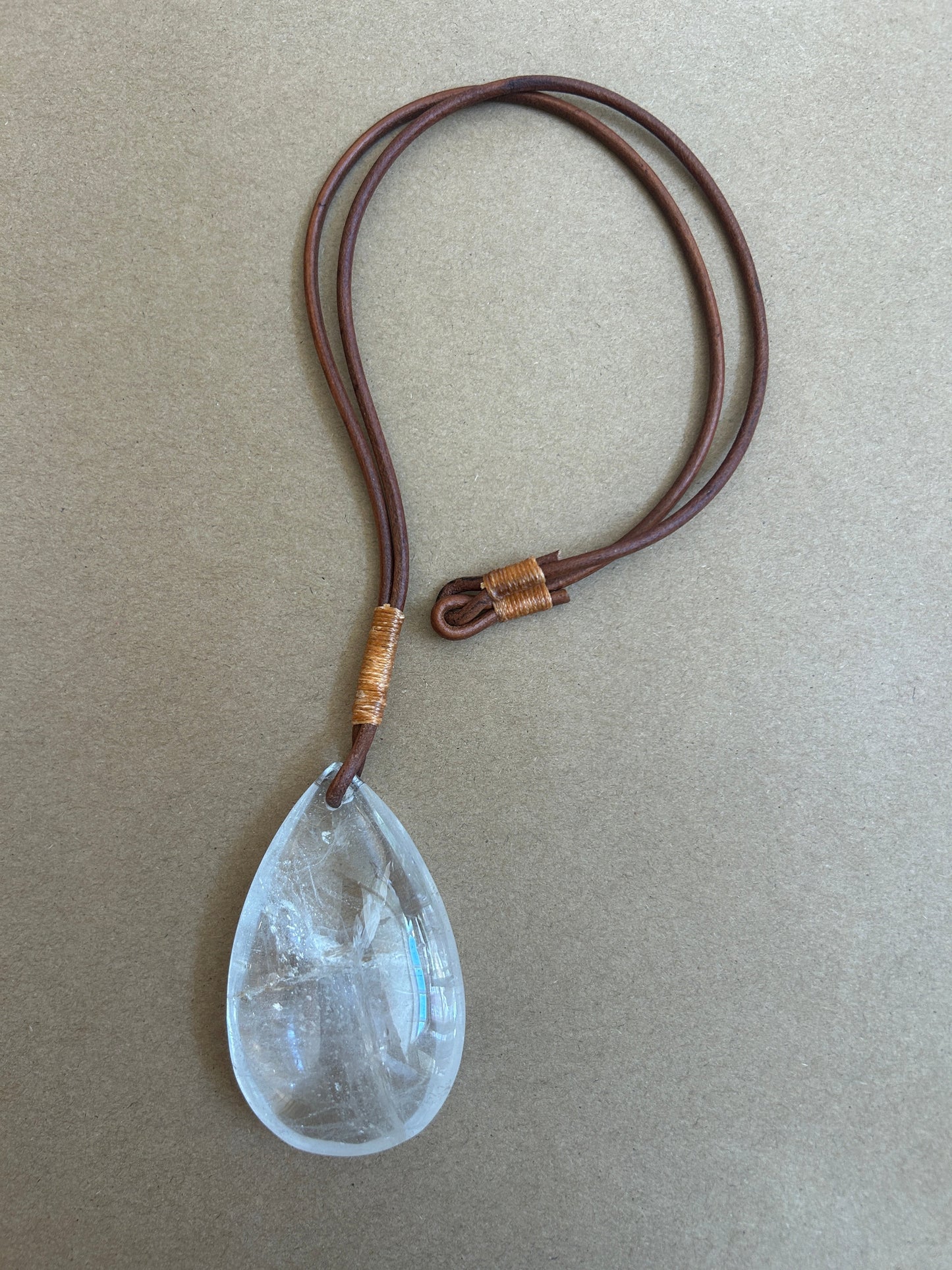 A teardrop-shaped clear crystal pendant necklace on a dark brown leather cord with gold-colored wrap accents, displayed on a textured beige background.