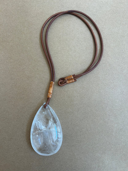 A teardrop-shaped clear crystal pendant necklace on a dark brown leather cord with gold-colored wrap accents, displayed on a textured beige background.