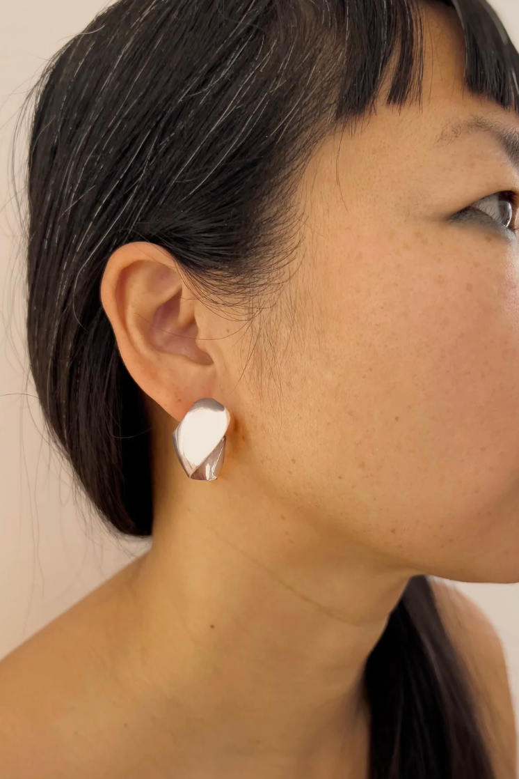 A close-up side profile of a woman wearing a single sculptural, polished silver earring on her ear.