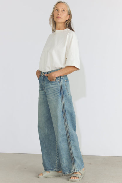 A woman modeling light-wash wide-leg denim jeans with a white short-sleeve top and gray slide sandals, standing against a plain white studio wall.