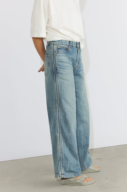 A person wearing light-wash wide-leg blue jeans, a white short-sleeve top, and beige sandals, standing against a plain white wall and concrete floor.