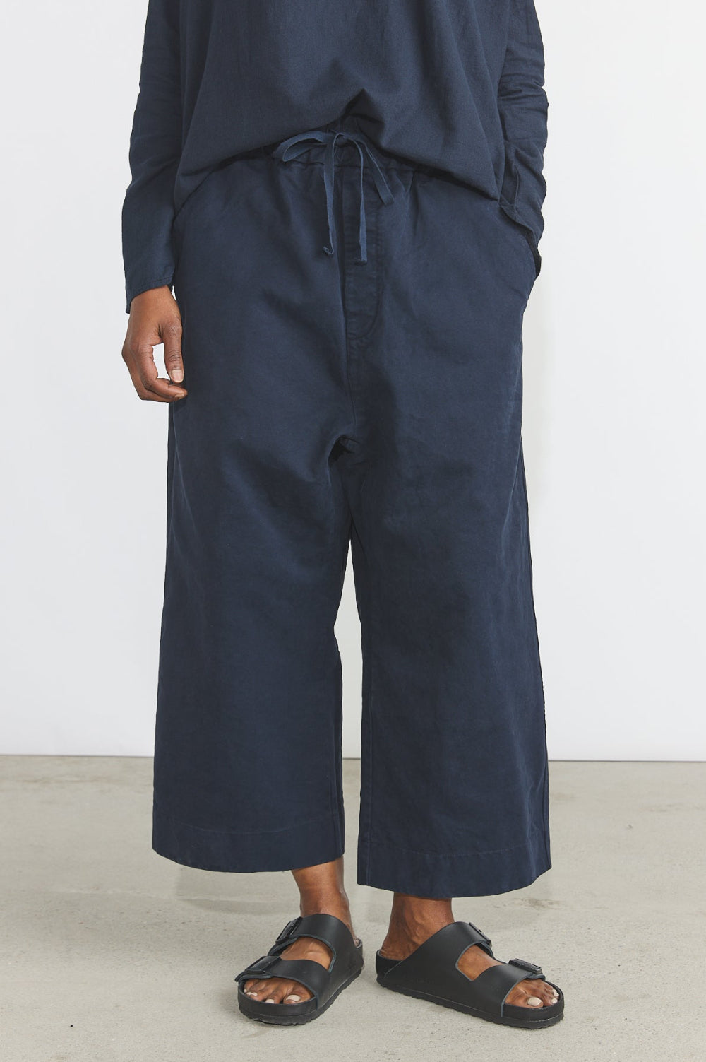 A person wearing loose dark navy wide-leg cropped pants with a drawstring waist, paired with a matching navy long-sleeve top and black slide sandals, standing against a plain light wall.