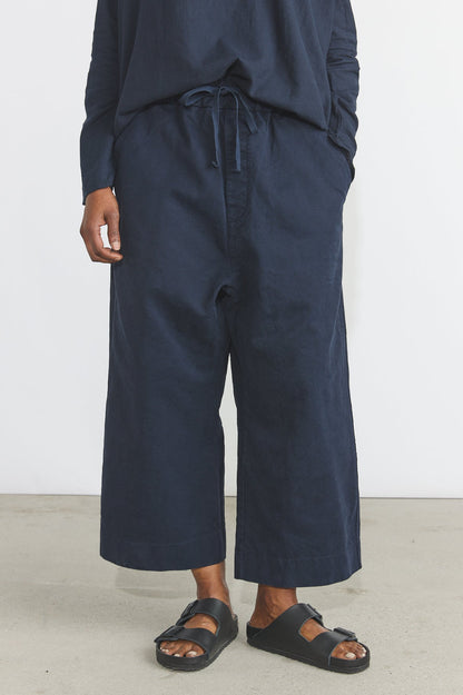A person wearing loose dark navy wide-leg cropped pants with a drawstring waist, paired with a matching navy long-sleeve top and black slide sandals, standing against a plain light wall.