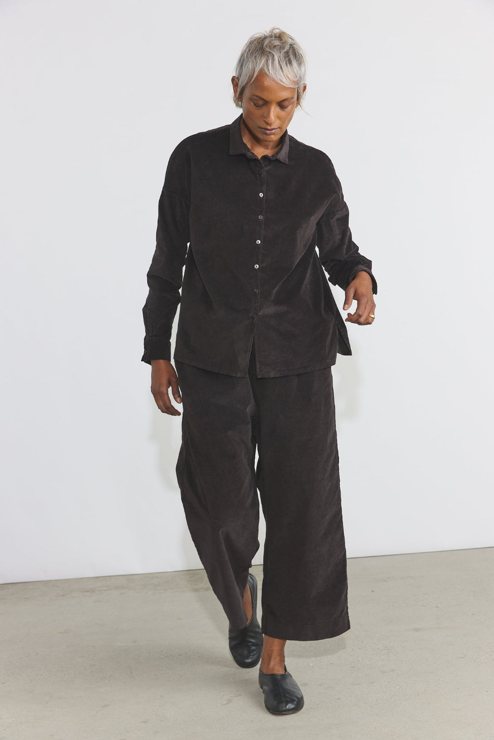 A model with short gray hair walks forward wearing a matching black corduroy button-up shirt and wide-leg cropped pants against a minimalist white studio background.