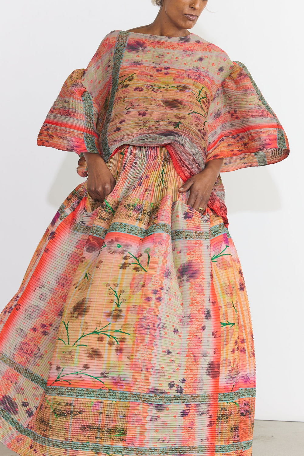 A model wearing a voluminous, wide-sleeved maxi dress with a vibrant orange, pink, and green floral pattern, photographed against a plain white studio background.