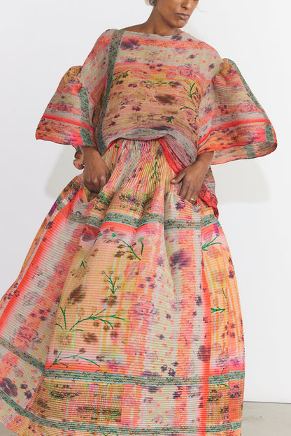 A model wearing a voluminous, wide-sleeved maxi dress with a vibrant orange, pink, and green floral pattern, photographed against a plain white studio background.