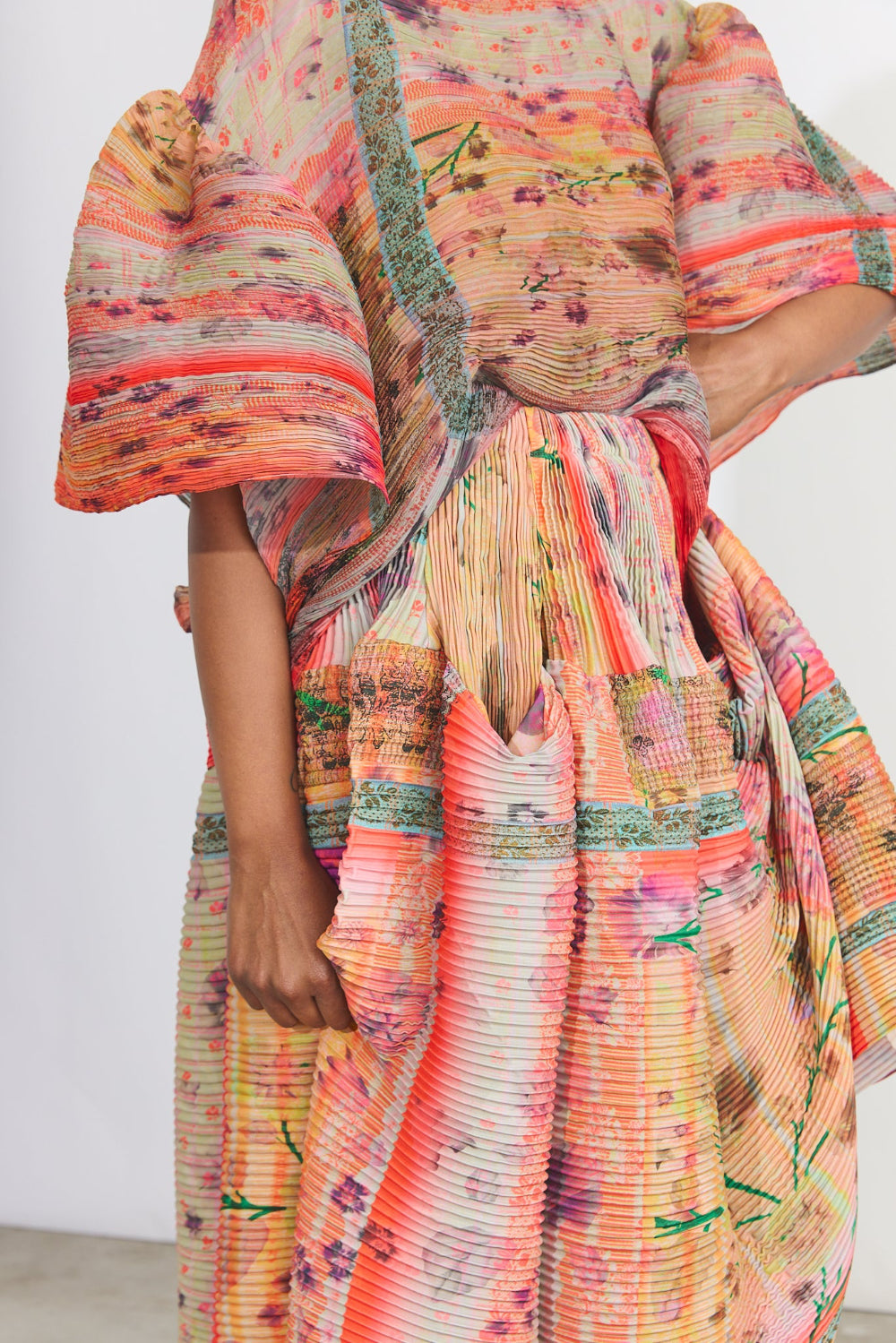 A close-up of a model wearing a pleated, multicolored dress with wide flared sleeves. The dress features a vibrant abstract pattern in shades of orange, pink, yellow, and green, with a pleated texture throughout.