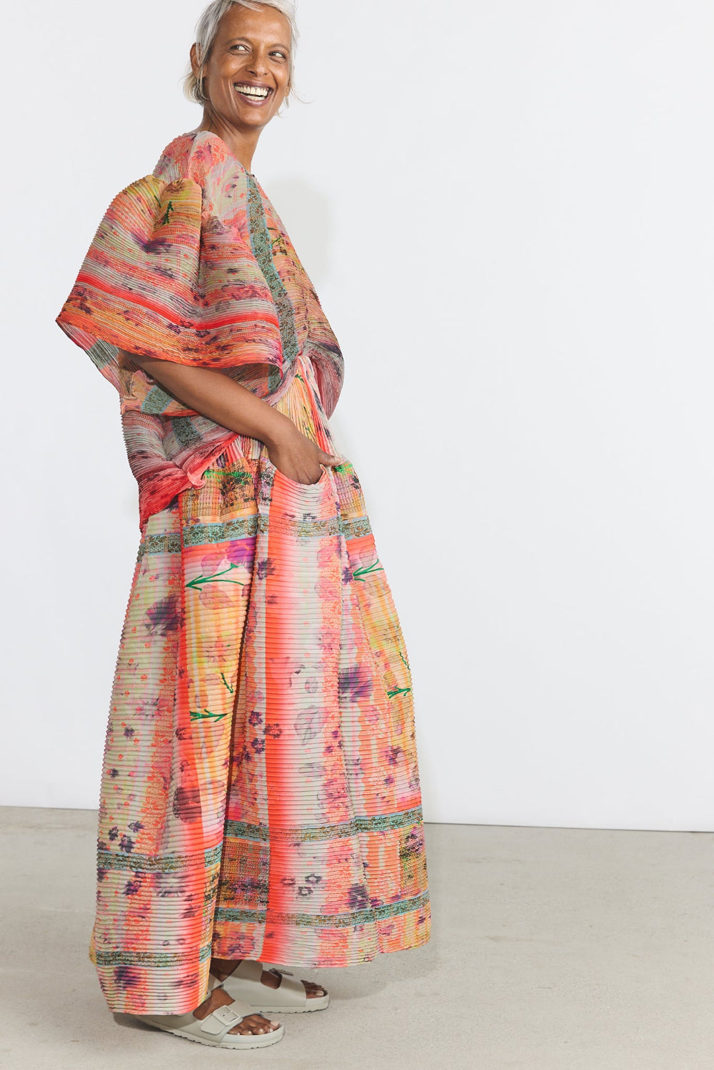 A woman modeling a loose, wide-sleeved maxi dress with a vibrant multicolor abstract print in shades of orange, pink, yellow, and green, set against a plain white studio background.