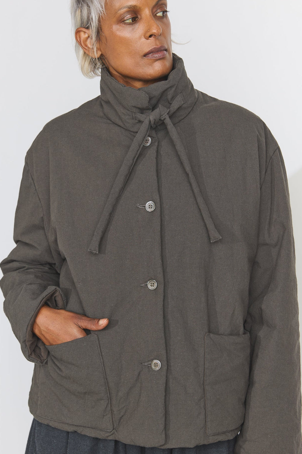 A model wearing a dark olive green quilted jacket with a high drawstring mock neck and front buttons, paired with a dark skirt, against a plain light gray background.