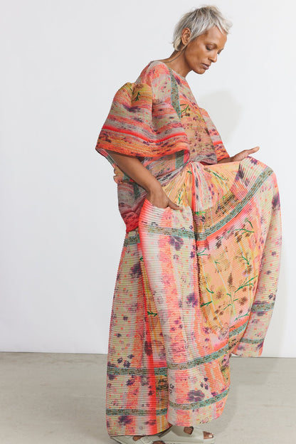 A woman wearing a loose, long-sleeved kaftan dress with a vibrant multicolor abstract print in shades of orange, pink, beige, and green, standing against a plain white studio wall.