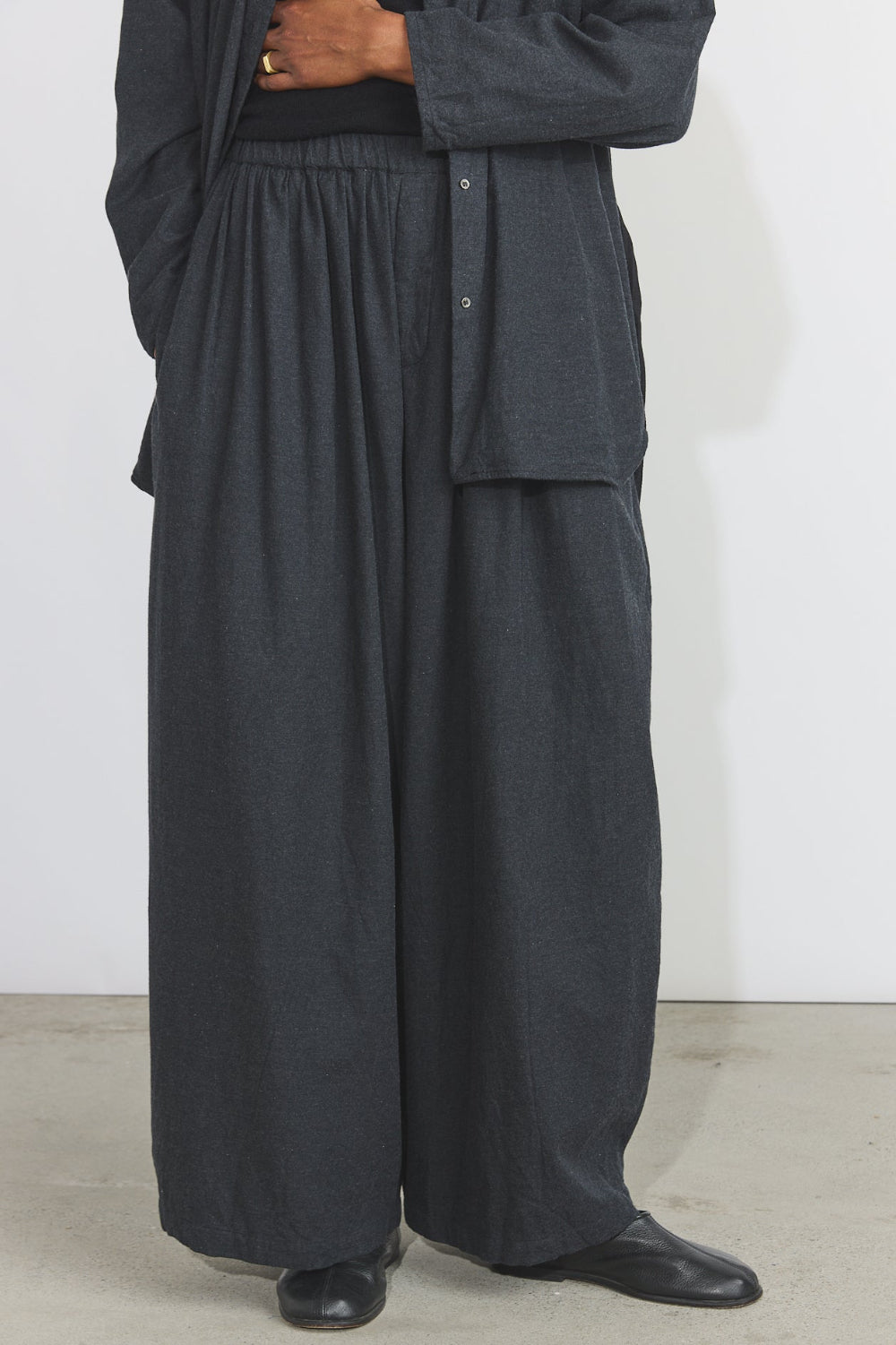 A person wearing loose, wide-leg dark charcoal gray pants with an elastic waistband, a matching dark button-front overshirt, and black shoes, standing against a plain light wall.