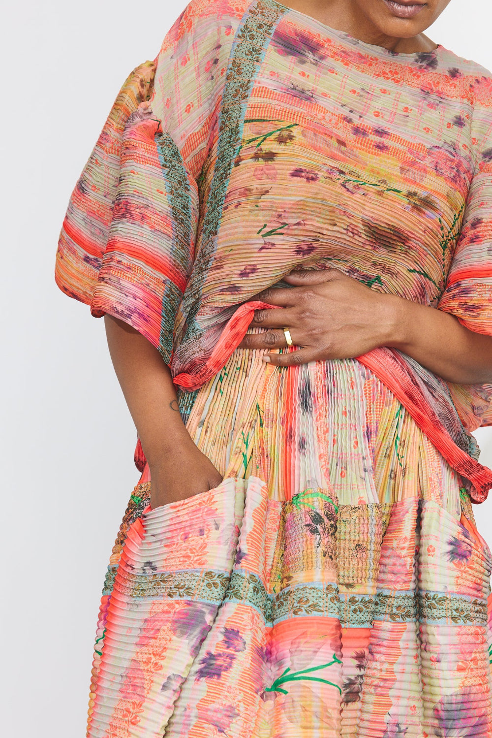 A close-up of a model wearing a pleated kaftan dress with a vibrant multicolor floral and striped pattern in shades of orange, pink, yellow, and green.