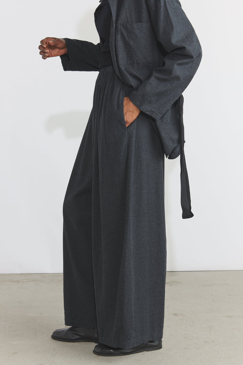 A person wearing a dark charcoal gray wide-leg jumpsuit or long robe-style pants with a self-tie belt, shown from the neck down against a plain light wall and concrete floor.