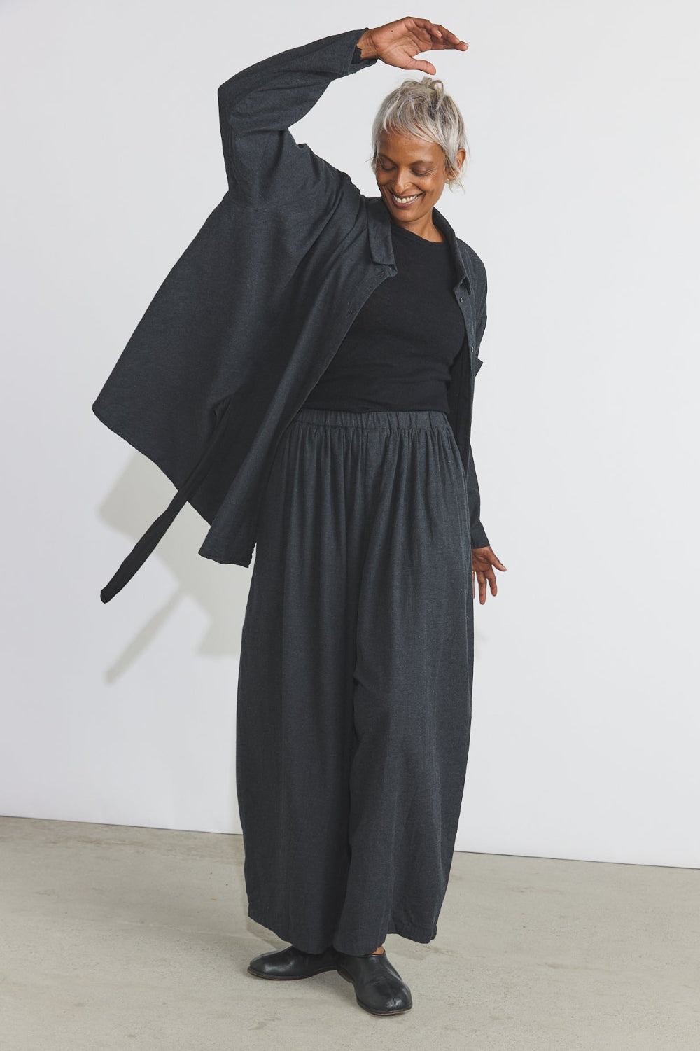 A model with short gray hair wearing a black long-sleeve top, wide-leg pleated trousers, and a draped black jacket, standing in a minimalist studio with a white wall and concrete floor.