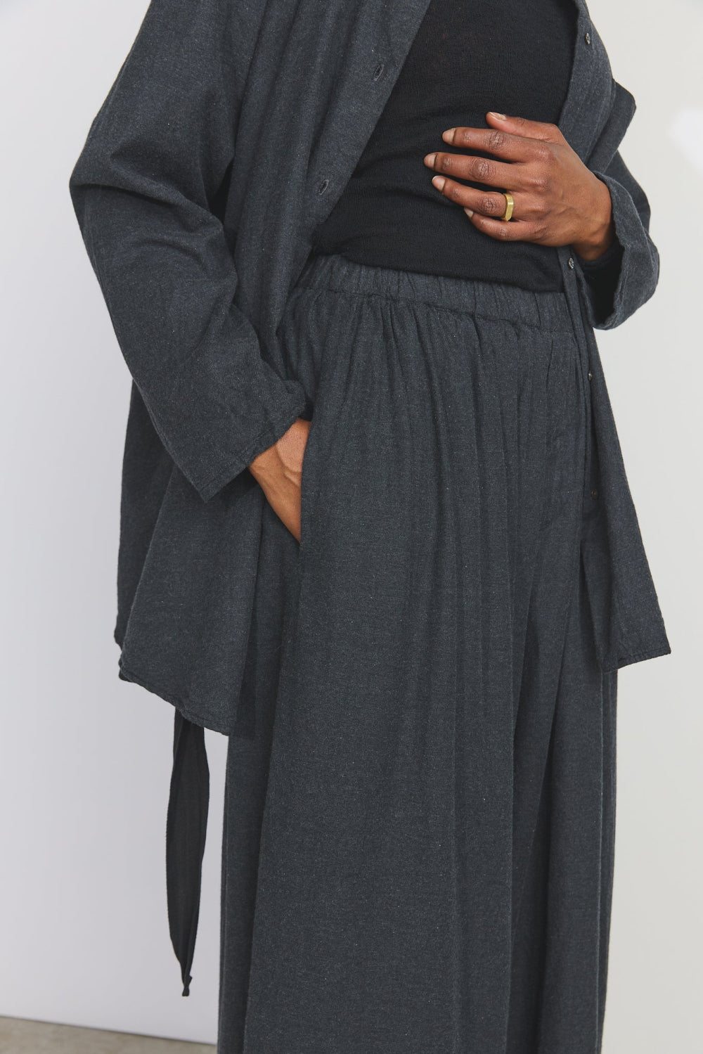 A person wearing a dark charcoal gray layered outfit, featuring a long-sleeve button-front cardigan over a black top and matching dark wide-leg trousers, shown from the chest down against a plain light gray background.