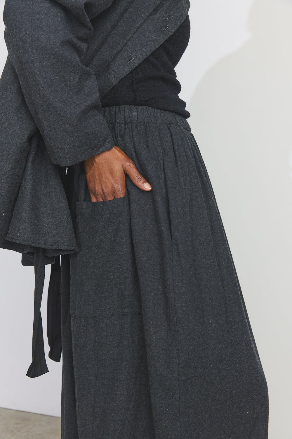 A close-up of a person wearing dark charcoal gray wide-leg trousers with side pockets, paired with a black top and draped dark gray outer layer, standing against a plain off-white wall.